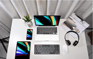 A KeyChron keyboard on a desktop with an Apple laptop, a tablet, a pair of headphones, and other tech.